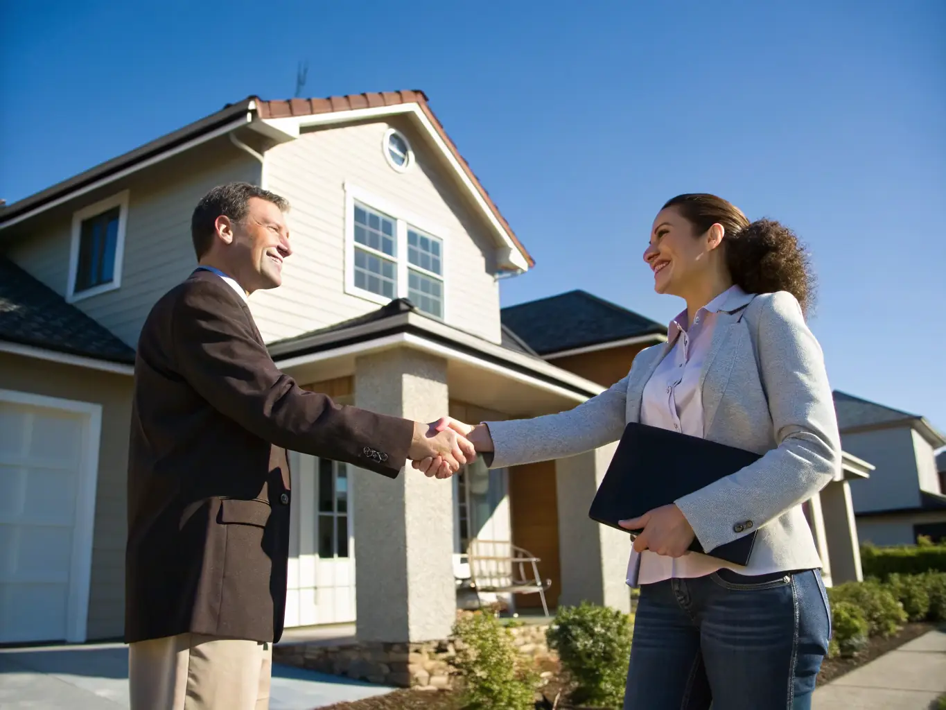 A professional real estate agent shaking hands with a satisfied client in front of a beautiful house. The image represents the success and growth potential offered through Agent Recruiting via REAL Broker.